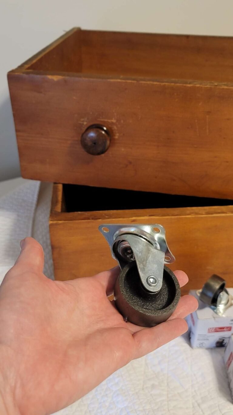 A hand holding a swivel caster wheel in the foreground. In the background, an open wooden drawer with a round wooden knob rests on a white quilted surface, offering easy under bed storage. Another caster wheel is partially visible next to the drawer.