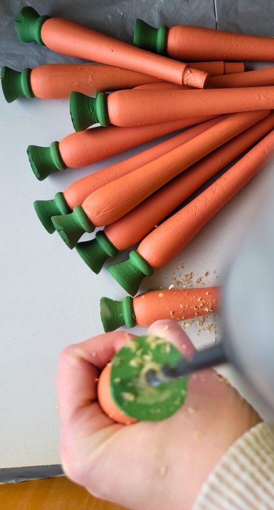 A hand is drilling into the green end of a wooden carrot, with wood shavings scattered around. This Carrot DIY project features several completed wooden carrots with orange bodies and green tops.