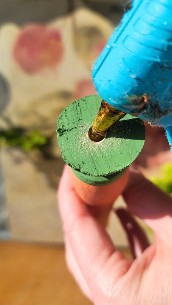 A close-up of a person using a blue hot glue gun to apply glue to the green top of a wooden spindle. The person's thumb and index finger hold the dowel steady, part of their ongoing spindle project.